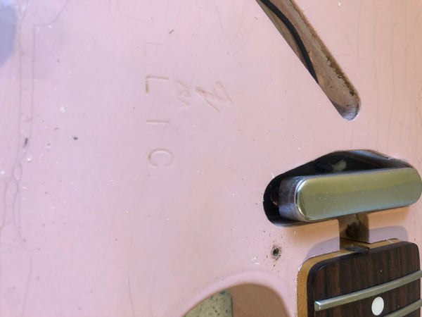 Close-up of pink electric guitar body showing faint serial number and part of the pickup and fretboard with dot inlay and metal fret