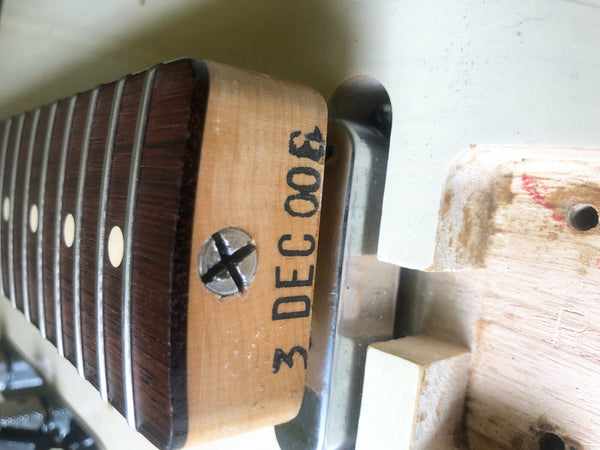 Close-up of guitar neck heel showing serial number 3 DEC 003 and fretboard with metal frets and dot inlays on a wooden electric guitar neck.