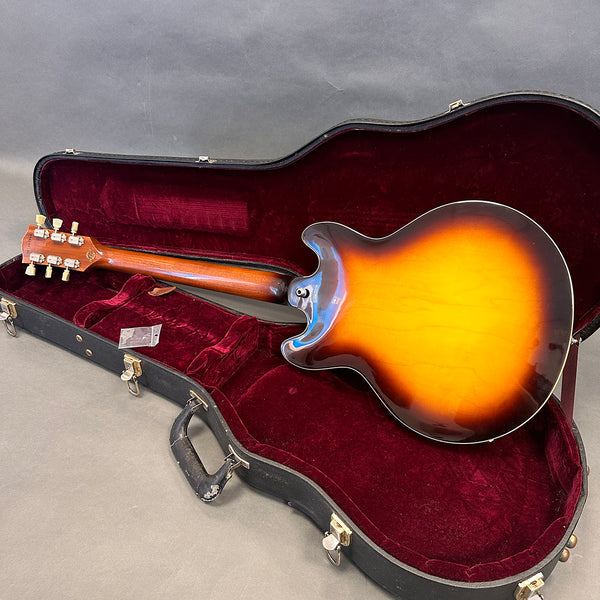 Sunburst semi-hollow electric guitar resting in open black hard case with red velvet interior, showing back and neck of guitar.