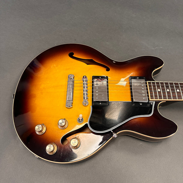 Semi-hollow body electric guitar with sunburst finish, two humbucker pickups, black pickguard, tune-o-matic bridge, and four control knobs on gray background