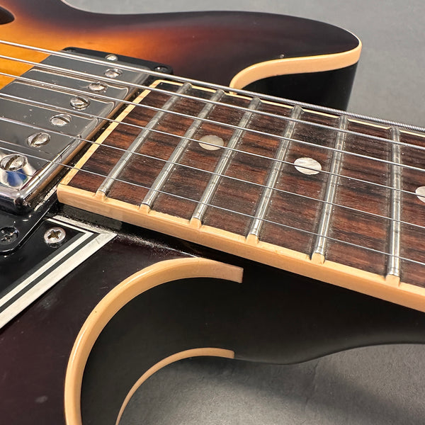 Close-up view of electric guitar neck and fretboard with metal frets and dot inlays, showing sunburst finish and chrome pickup with strings over semi-hollow guitar body.