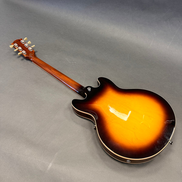 Back view of sunburst semi-hollow electric guitar with wooden neck and vintage tuning pegs resting on gray surface