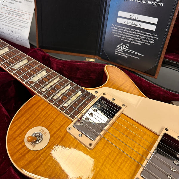 Close-up of Gibson Les Paul electric guitar with honey burst finish, cream pickguard, humbucker pickups, toggle switch, and certificate of authenticity in velvet-lined guitar case