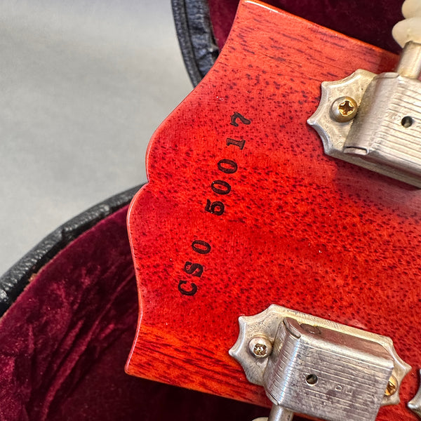 Close-up of guitar headstock with serial number CS0 50017 stamped on reddish wood near vintage metal tuning pegs in guitar case lined with red velvet
