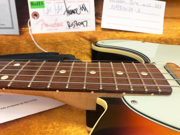 Close-up of Fender electric guitar rosewood fretboard and strings with sunburst finish body and white pickguard, displayed with Fender certification tags in the background on orange plush case lining.