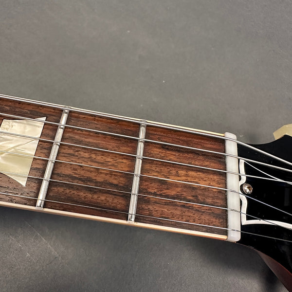 Close-up of electric guitar fretboard showing frets, strings, and inlay on dark wood fingerboard with black headstock against gray background