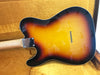 Sunburst finish electric guitar body with maple neck and chrome neck plate, resting on a mustard yellow plush surface, showcasing the rear side with string ferrules visible.