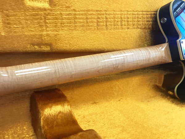 Close-up of a glossy figured maple guitar neck resting on a yellow plush guitar case interior with a dark blue guitar body partially visible.