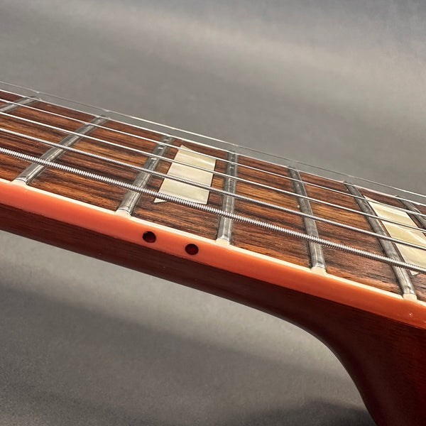 Close-up of guitar neck and fretboard showing metal frets, pearl inlay markers, and wound steel strings on a wooden guitar neck with orange binding