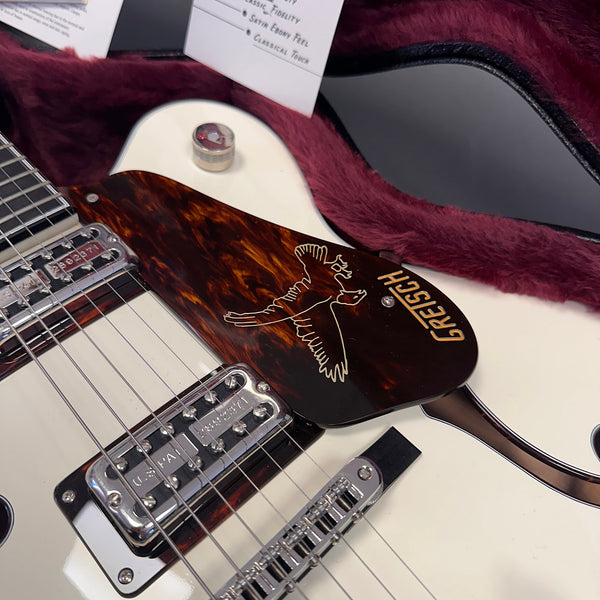 Close-up of white Gretsch electric guitar with tortoiseshell pickguard featuring a golden bird design and Gretsch logo, in a plush purple-lined guitar case.