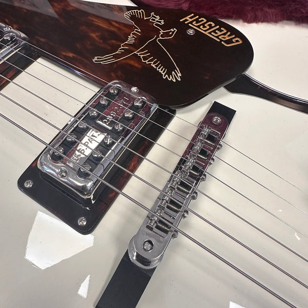 Close-up of Gretsch electric guitar bridge and pickup with bird logo on pickguard, white guitar body with chrome hardware and six strings.