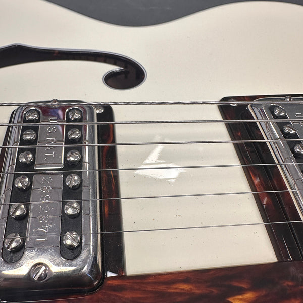 Close-up of white electric guitar body with metal humbucker pickups and strings, showing f-hole and tortoiseshell trim
