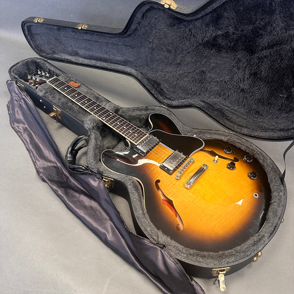 Sunburst semi-hollow electric guitar with double cutaway body in black plush-lined hardshell case and black gig bag side by side on gray surface