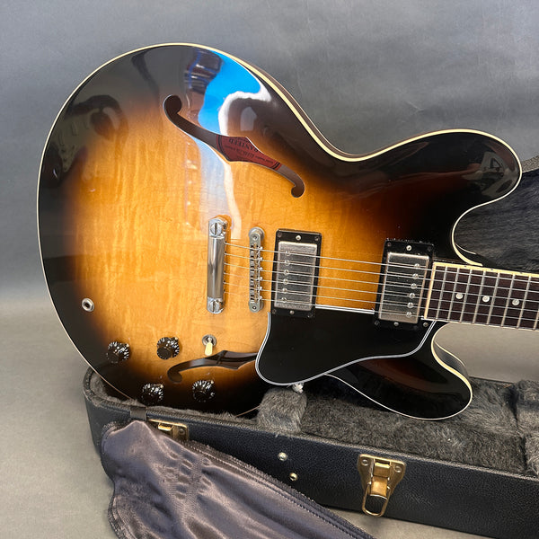 Sunburst semi-hollow electric guitar with dual humbucker pickups, black pickguard, and control knobs, resting in an open black hard case with plush interior lining.