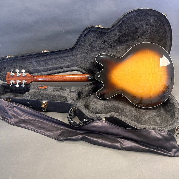 Electric semi-hollow body guitar with sunburst finish lying face down in open black hard case with gray plush interior and soft cover on gray surface