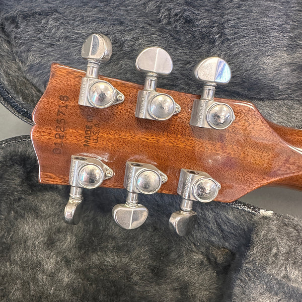 Close-up of mahogany guitar headstock with Grover tuning pegs and visible serial number and "Made in USA" stamp, resting on a plush gray guitar case lining