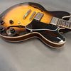 Semi-hollow body electric guitar with sunburst finish, dual humbucker pickups, black pickguard, and cream binding on grey background
