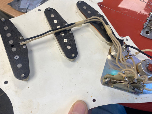Close-up image of vintage single coil pickups and wiring on a white guitar pickguard, showing aged cloth-covered wires and potentiometers for electric guitar electronics repairs and restoration.