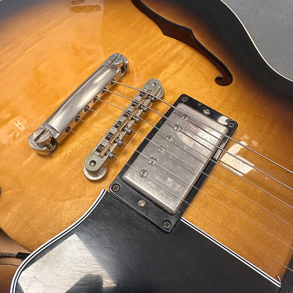 Close-up of vintage semi-hollow electric guitar body showing sunburst finish, chrome bridge, strings, and black pickguard with white trim.