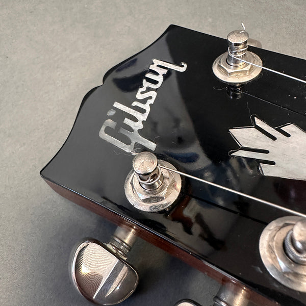Close-up of black Gibson guitar headstock with silver tuning pegs and inlaid logo on grey background