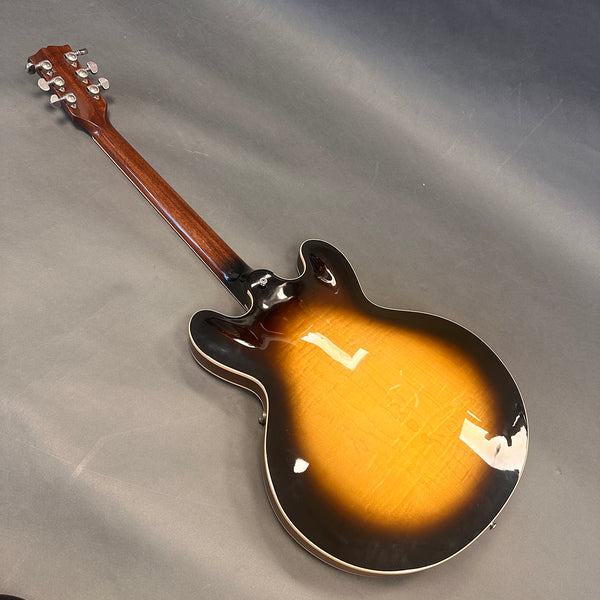 Back view of a sunburst semi-hollow electric guitar with a wooden neck and chrome tuning pegs against a gray background