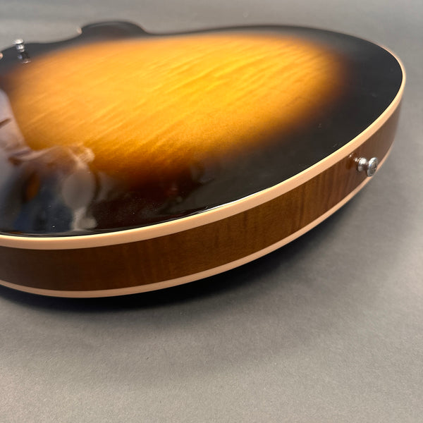 Close-up of the edge and strap button of a sunburst finish acoustic guitar body with a wooden side and cream binding on a gray background