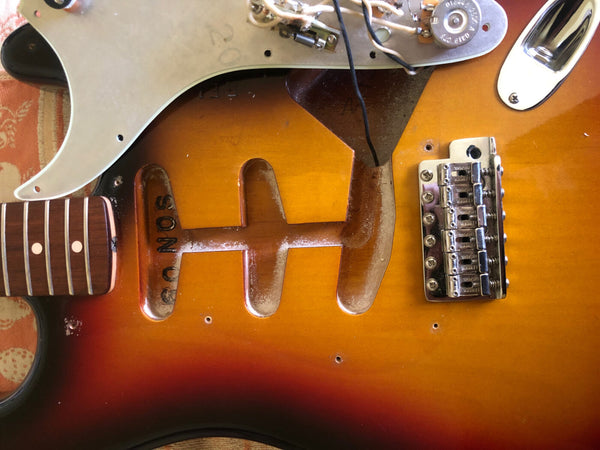 Close-up of vintage sunburst electric guitar body with pickguard and exposed pickup cavity, showing wood grain and chrome bridge saddle detail.