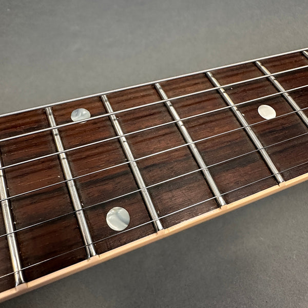 Close-up of rosewood guitar fretboard with metal frets, mother-of-pearl dot inlays, and steel guitar strings on light maple neck edge against gray background