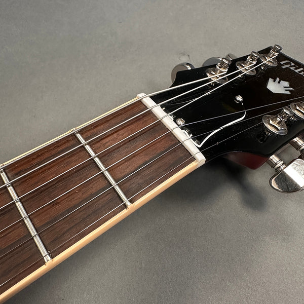 Close-up of guitar neck, rosewood fretboard, metal frets, and black headstock with tuning pegs and white nut on gray background