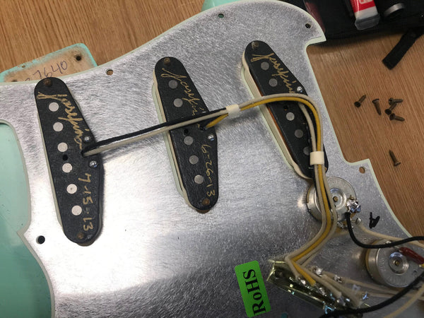 Close-up of vintage Fender guitar pickups mounted on a metallic pickguard with wiring and potentiometers, showing hand-written dates on pickups and a green RoHS compliance sticker, laid on a wooden surface with small screws nearby