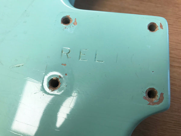 Close-up of the back of a seafoam green guitar neck with visible screw holes and the word "RELIC" embossed in the finish showing natural wear and chipping around the edges.