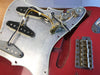 Close-up of vintage electric guitar with red body, exposed pickguard wiring, three single-coil pickups, two control knobs, and chrome bridge plate with individual adjustable saddles.