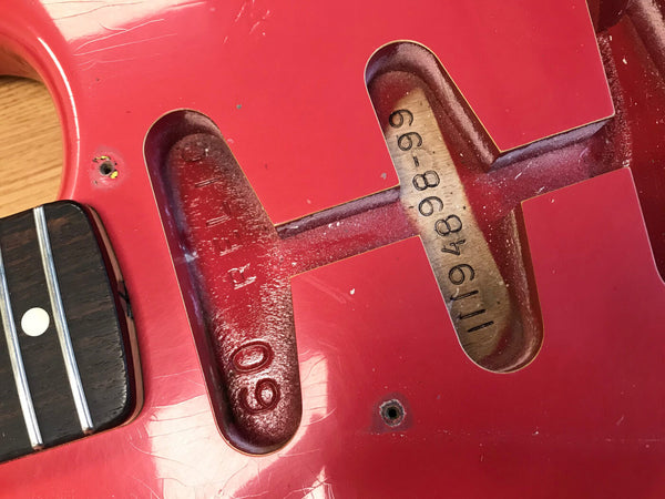 Close-up of red electric guitar body without pickups, showing neck pocket with serial number 111948899 and 60 relic stamp inside pickup cavity near rosewood fingerboard and metal frets