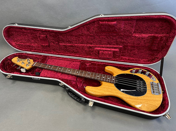 Natural wood finish electric bass guitar with black pickguard and chrome tuning pegs in open red velvet-lined hard case on gray background