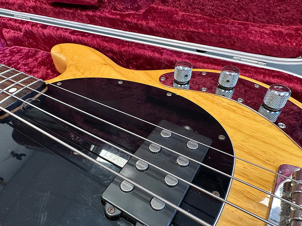 Close-up of natural wood electric bass guitar body with black pickguard, metal control knobs, and four strings in a red plush guitar case