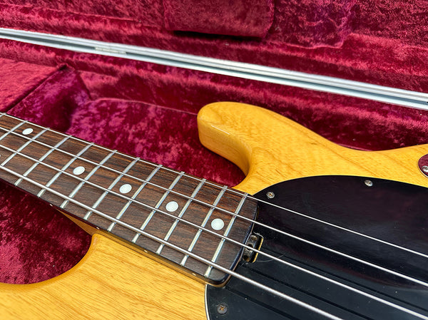 Close-up of natural wood electric bass guitar neck and fretboard with white dot inlays and black pickguard inside red velvet guitar case