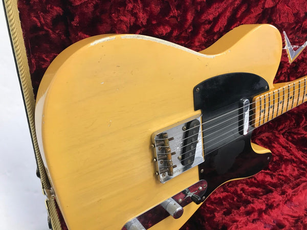 Close-up of vintage blonde electric guitar body with black pickguard, single-coil pickups, and chrome bridge hardware, resting in red velvet-lined case with tweed exterior strap visible.