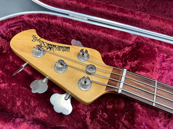 Close-up of a Music Man StingRay electric bass guitar headstock with tuning pegs and strings, resting in a red velvet guitar case.