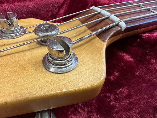 Close-up image of tuning pegs and strings on a wooden bass guitar headstock with a red velvet background, showcasing metal hardware and string winding details.