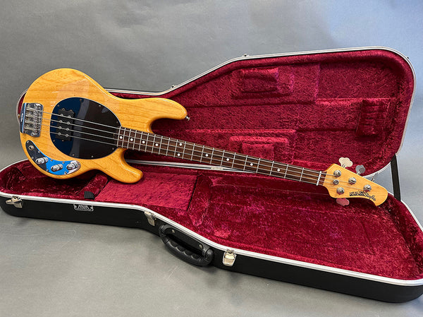 Natural finish Music Man StingRay bass guitar with black pickguard inside red velvet-lined hard case on gray surface