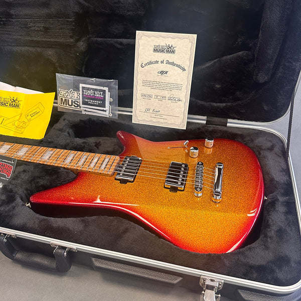 Ernie Ball Music Man electric guitar in sparkly red-orange finish with dual humbucker pickups inside a black plush-lined hard case with certificate of authenticity and accessories.