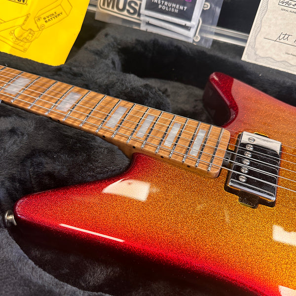 Close-up of electric guitar with red and orange sparkle finish, maple fretboard with rectangular inlays, resting in black plush-lined guitar case with instrument polish and paperwork visible.