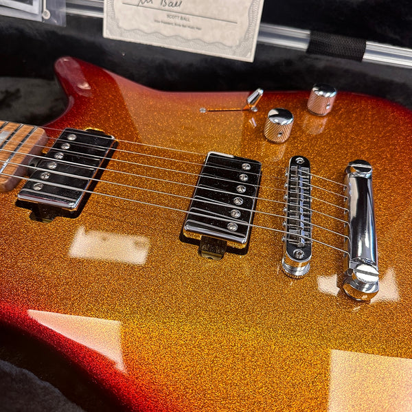 Close-up of electric guitar body with gold and red sparkle finish, chrome bridge, two humbucker pickups, and control knobs, displayed in black plush case with certificate of authenticity.