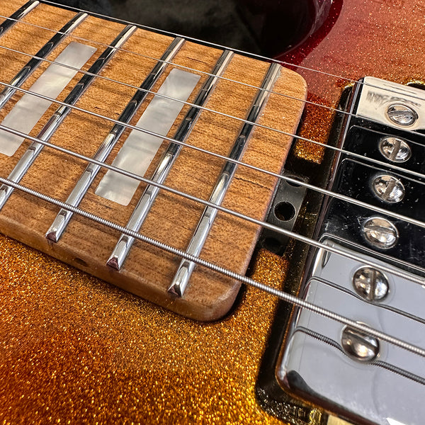 Close-up of electric guitar fretboard with rectangular inlays and metal frets, featuring a metallic orange glitter finish and chrome pickups and strings.
