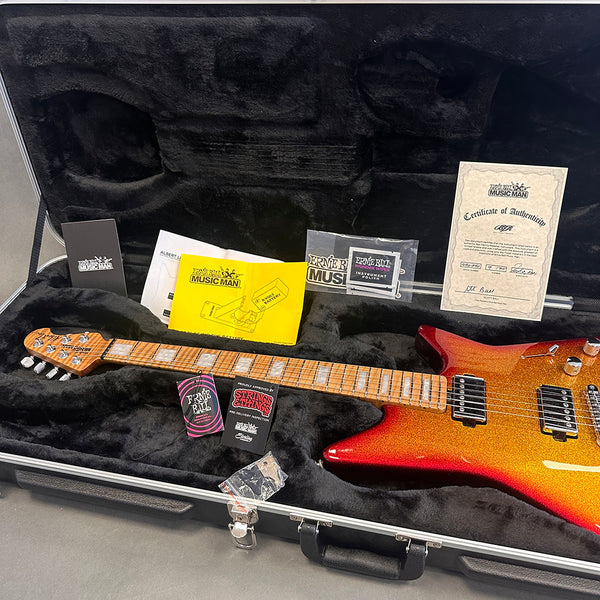 Ernie Ball Music Man signature electric guitar with flamed maple neck and sunburst orange sparkle finish in black plush-lined hard case with certificate of authenticity and accessory documentation.