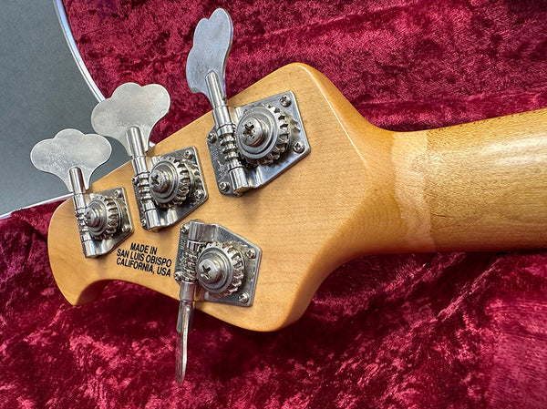 Close-up of bass guitar headstock tuning pegs with "Made in San Luis Obispo California, USA" stamp on maple neck, lying on red velvet fabric.