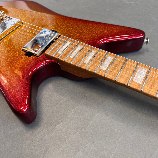Close-up of electric guitar with sparkly red and orange finish and maple fretboard with rectangular inlays on gray surface