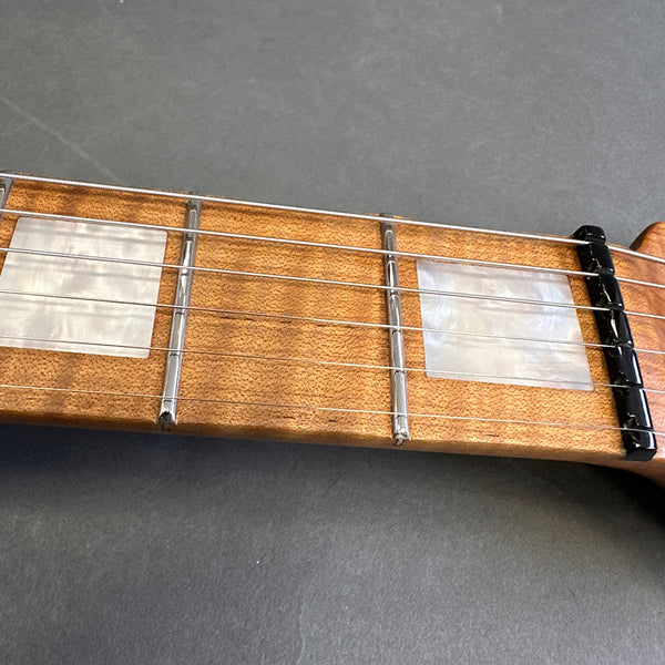 Close-up of guitar fretboard with mother-of-pearl square inlays and metal frets on a light wooden neck with steel strings and black nut.