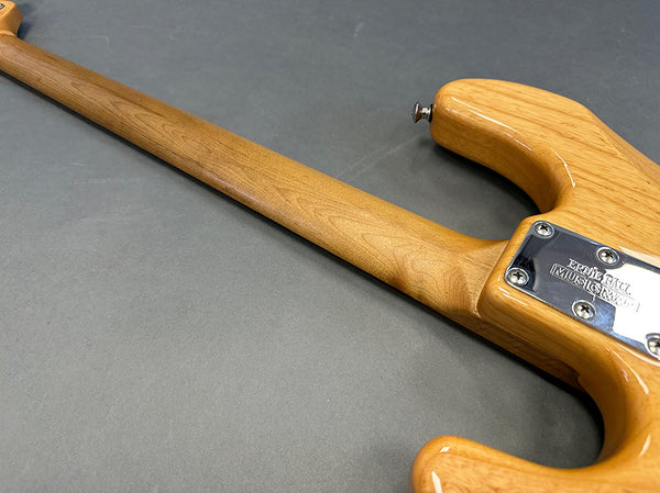Close-up of natural wood grain neck and back of electric guitar body with metal neck plate engraved "Ernie Ball Music Man" on gray background