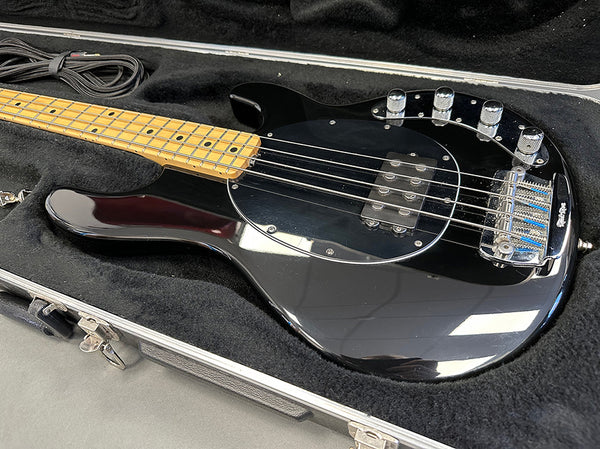 Black electric bass guitar with maple neck and four control knobs in open black hard case on gray surface